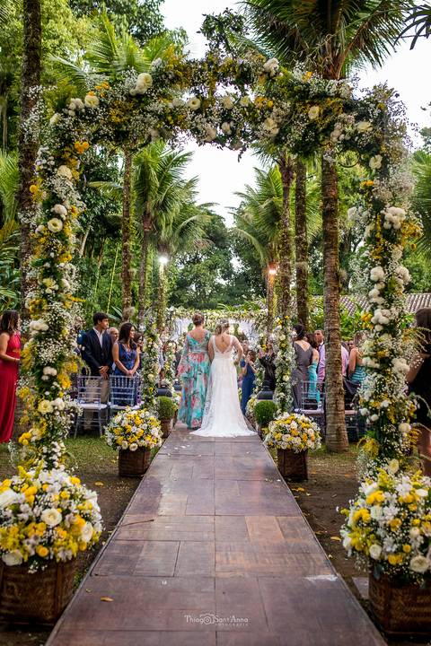 entrada da noiva com sua mãe decoração vegetal convidados observam de pé flores brancas e amarelas '