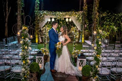 noivos se abraçam decoração altar ao fundo flores amarelas e brancas cadeiras brancas para convidados '