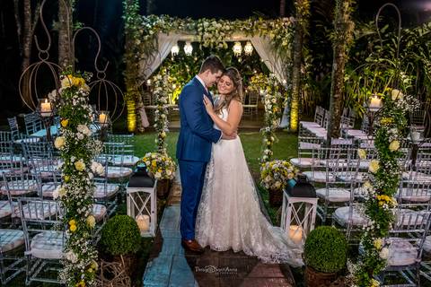 noivos se abraçam decoração altar ao fundo flores amarelas e brancas cadeiras brancas para convidados '