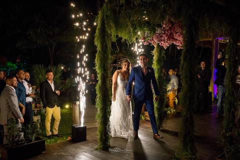 noivos atravessam portal de madeira revestido de vegetação com flores cor de rosa '