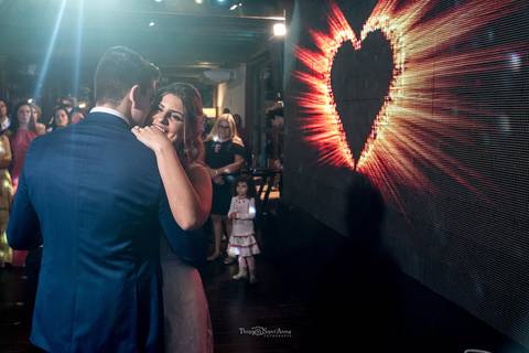 noivos dançam valsa em frente a telão com imagem de coração convidados observam '