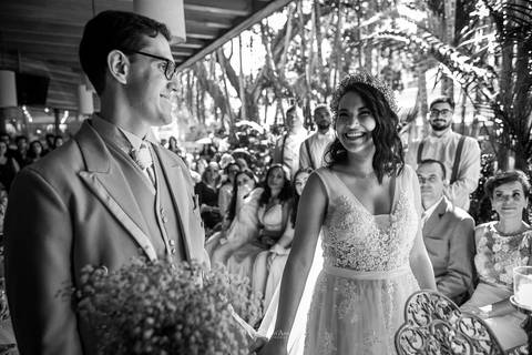 noivos de mãos dadas a noiva sorri convidados ao fundo preto e branco'