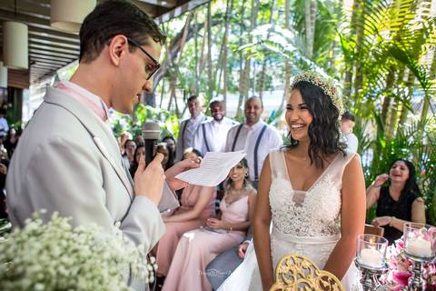 noivo lê seus votos de casamento noiva sorri padrinhos observam sorrindo'