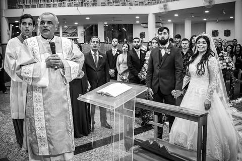 noivos e padre no altar noiva sorri convidados ao fundo preto e branco'