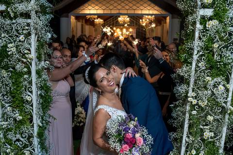 noiva sorri buque tons de rosa lilás noivo a abraça decoração arco vegetal flores brancas padrinhos ao fundo'