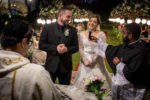 noiva sorri falando ao microfone noivo sorri na altar junto ao padre'