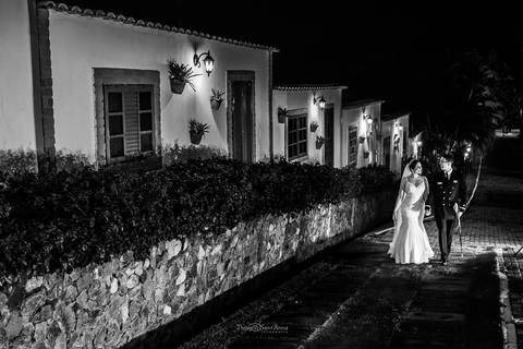 noiva e noivo caminham por rua de paralelo ao lado de casas rústicas preto e branco'