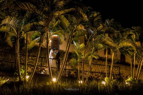 coqueiros iluminados noiva e noivo se abraçam casa rústica ao fundo'