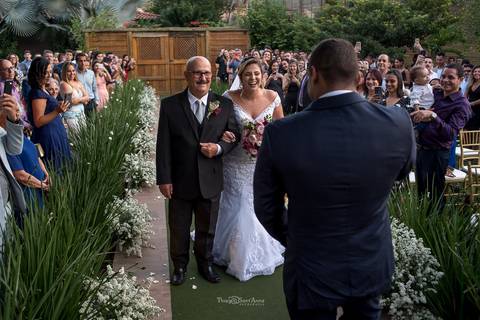 noiva e seu pai sorrindo ao se aproximar do noivo decoração vegetação flores brancas convidados observam de pé'