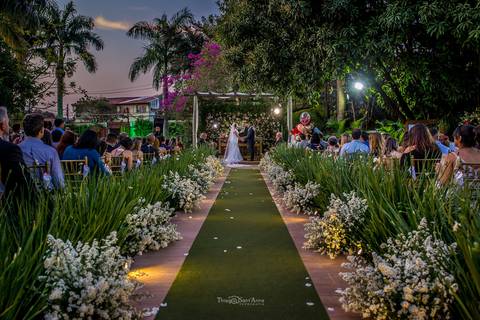 noivos no altar tenda madeira tecido branco convidados observam vegetação balões flores tapete verde'