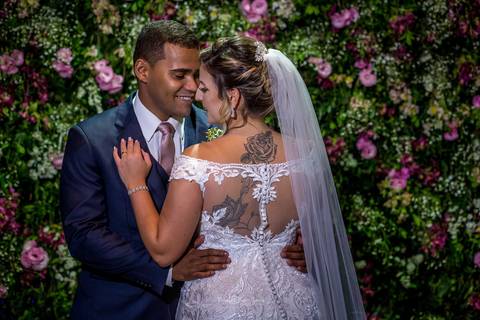 noivos se abraçam noiva vestido véu parede flores tons de rosa ao fundo'