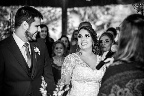 noiva observa noivo sorri convidados ao fundo preto e branco'