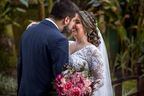 noivos se abraçam noiva sorri buque flores tons de rosa vegetação ao fundo'