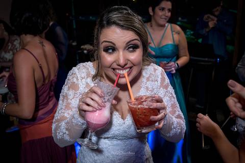 noiva toma dois drinks de canudinho sorri convidadas ao fundo'