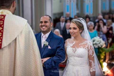 noivos sorrindo no altar de frente ao padre convidados ao fundo'