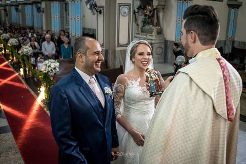 noivos sorrindo junto ao padre igreja convidados ao fundo decoração flores brancas'