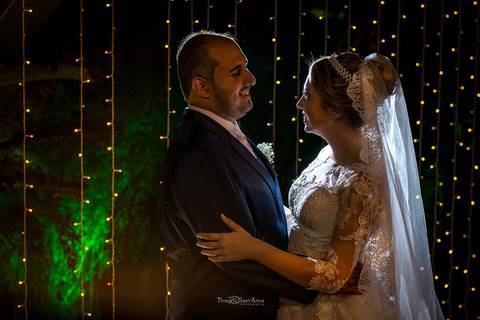 noivos se abraçam sorrindo vegetação ao fundo luzes decorativas'