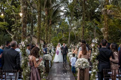 entrada da noiva com seu pai convidados observam de pé ambiente externo vegetação coqueiros decoração flores brancas'
