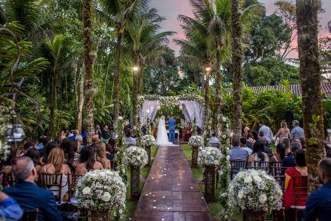 noivos no altar ambiente externo vegetação coqueiros flores brancas convidados observam'