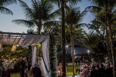 ambiente externo vegetação coqueiros tenda madeira tecido branco noivos no altar convidados observam'