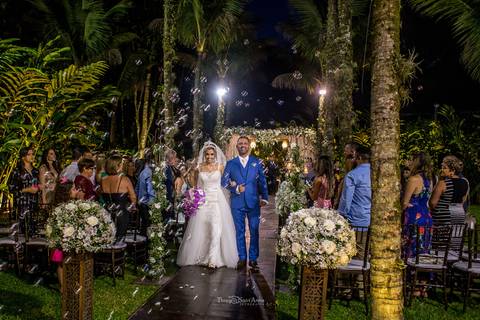 saída dos noivos noiva vestido véu buque em tons de rosa decoração vegetação flores brancas convidados observam de pé'