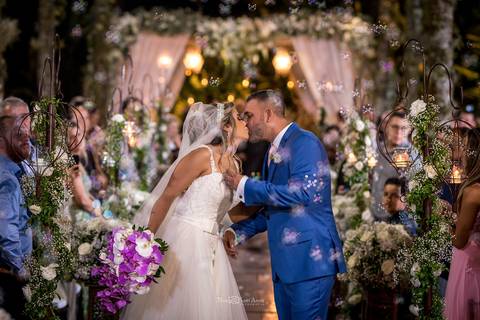 beijo dos noivos noiva vestido véu buque em tons de rosa decoração vegetação flores brancas convidados observam de pé'