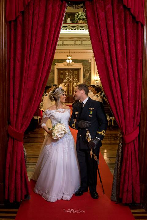 noivos se observam sorrindo noiva vestido véu buque flores brancas cortina vermelha'