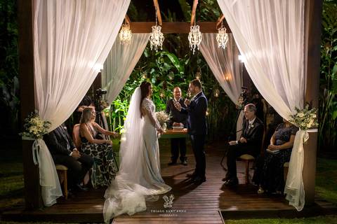 noivos altar madeira tecidos brancos luminárias familiares observa cerimônia'