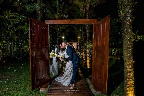beijo dos noivos em arco de porta madeira ambiente externo vegetação coqueiros'
