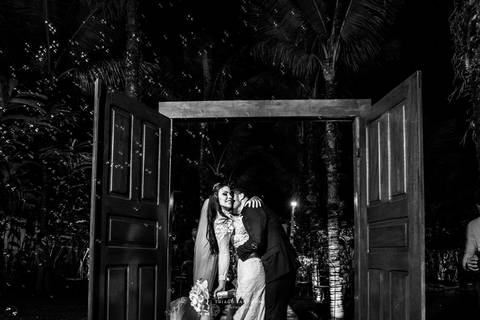 beijo dos noivos em arco de porta madeira ambiente externo vegetação coqueiros preto e branco'