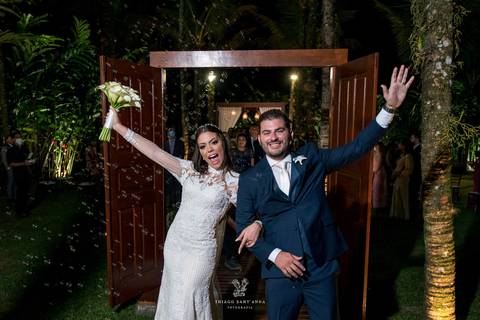 noivos sorrindo estendem os braços para o alto ao fundo um arco de portas madeira vegetação coqueiros'