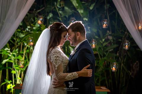 noivos posam abraçados noiva vestido véu noivo terno azul vegetação velas ao fundo velas'