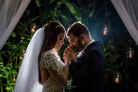 noivos posam abraçados noiva vestido véu noivo terno azul vegetação velas ao fundo velas'