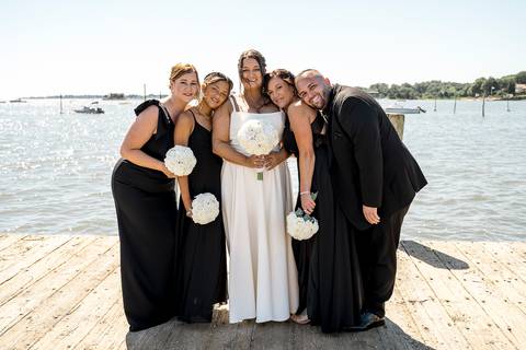 Group shot of the bridal party standing in a row by the water at Stony Creek, holding bouquets and smiling with joy.'