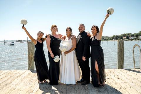 Group shot of the bridal party standing in a row by the water at Stony Creek, holding bouquets and smiling with joy.'