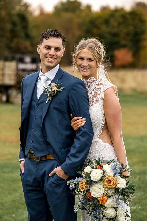 Cara and Daniel pose in front of a backdrop of fiery fall foliage at StoneHurst, their smiles glowing as they hold hands, framed by nature’s colorful embrace on a perfect autumn day.'