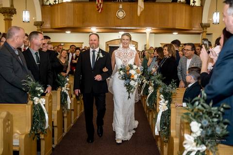 The newlyweds share a romantic first dance under twinkling lights at StoneHurst’s rustic barn, surrounded by cheering guests, while warm autumn colors fill the space with a cozy, festive atmosphere.'