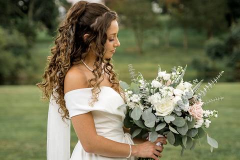 The bride stands in her elegant white gown, her curly hair framing her face beautifully as she holds a blooming bouquet of white and pink flowers'