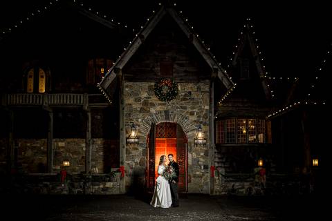 The newlyweds pose beneath the castle’s archway, surrounded by a halo of warm light against the cold night air, capturing the dramatic beauty of their winter wedding.'