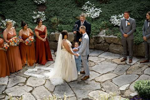 The bride walks down the aisle between her proud parents, sunlight pouring over the Heritage Hotel garden in Southbury, CT — pure elegance and emotion.'
