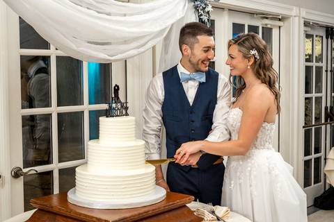The couple shares a sweet laugh while cutting their elegant white wedding cake, surrounded by loved ones.'