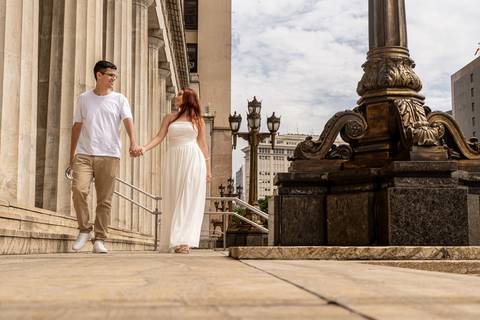 Júlia e Leonardo caminham de mãos dadas junto à arquitetura histórica do Centro do Rio de Janeiro.'