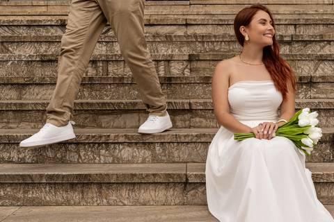 Júlia sentada em uma escadaria com flores enquanto Leonardo caminha durante o ensaio pré casamento.'
