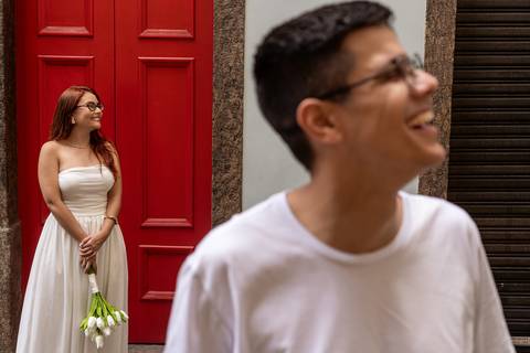 Júlia diante de uma porta vermelha e Leonardo sorrindo em primeiro plano durante ensaio pré casamento.'