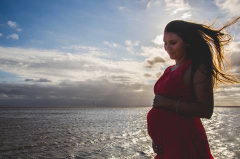mamãe com a mão no barrigão de vestido vermelho e o mar de  fundo no click da fotógrafa de gestante e família Keiti Santos em linda imagem'