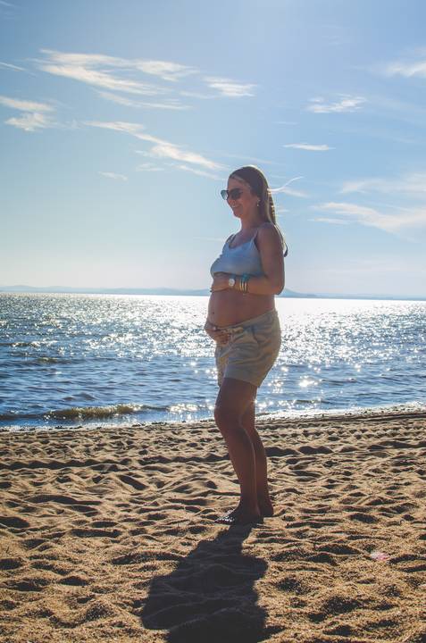 Mamãe Geórgia segurando sua linda barriga, com um lindo rio atrás e um sol brilhando no céu azul.'