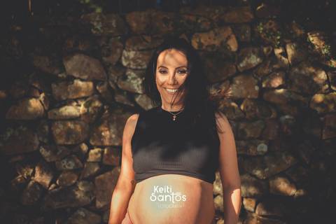 Mulher gestante sorrindo, ao fundo muro de pedras rústicas '