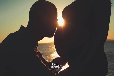 Homem beijando a barriga da esposa, imagem em contra luz, sol brilhando ao fundo'