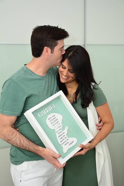 Imagem colorida, onde o casal segura um quadro com o nome Gabriel e uma oração'
