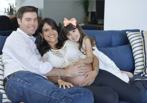 Imagem colorida, onde o casal e sua filha estão abraçados, sentados num sofá azul marinho'